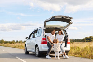 Outdoor shot of happy smiling woman and man drinking hot tea from thermos flask sitting in car trunk with their baby daughter, wife and husband spending good time together while traveling.